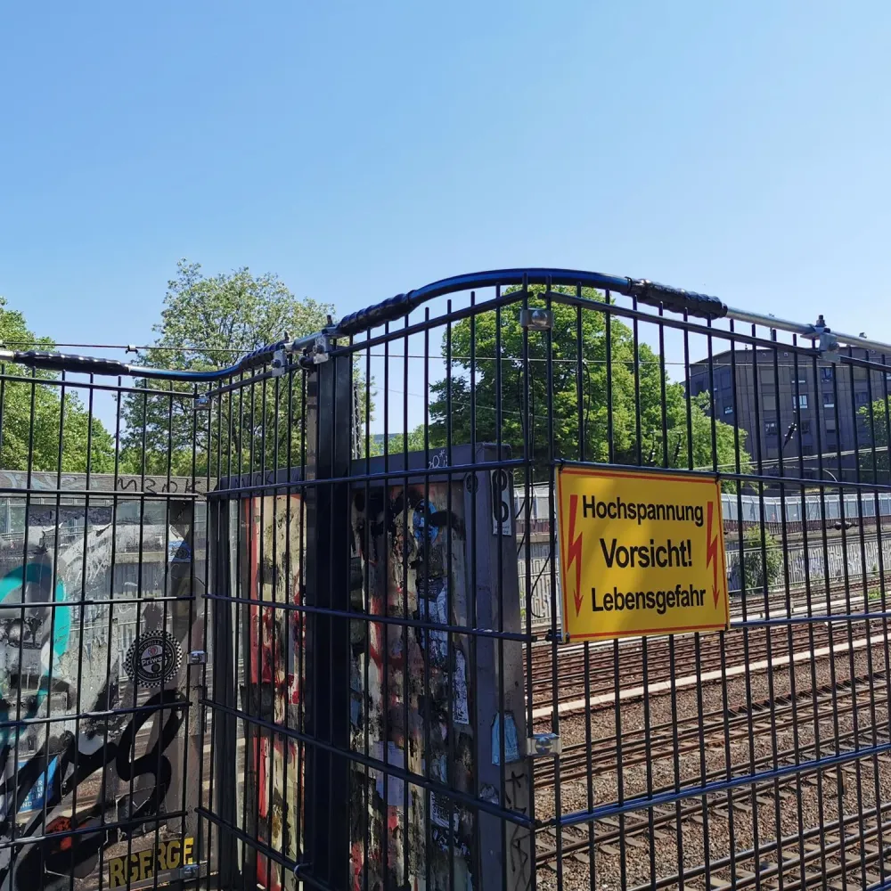 Fenced railway area with a yellow high-voltage warning sign under a clear blue sky.