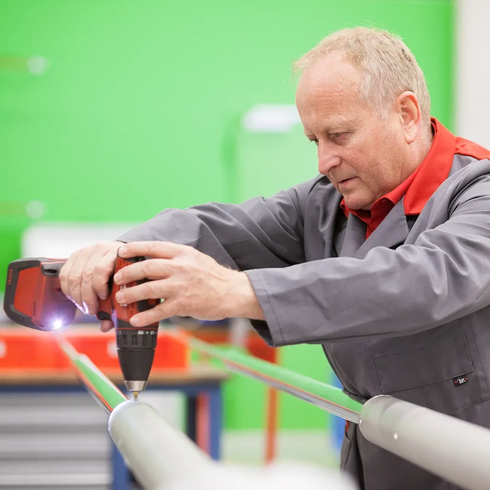 Worker in a grey lab coat using a cordless drill to fasten a metal rod in a workshop.