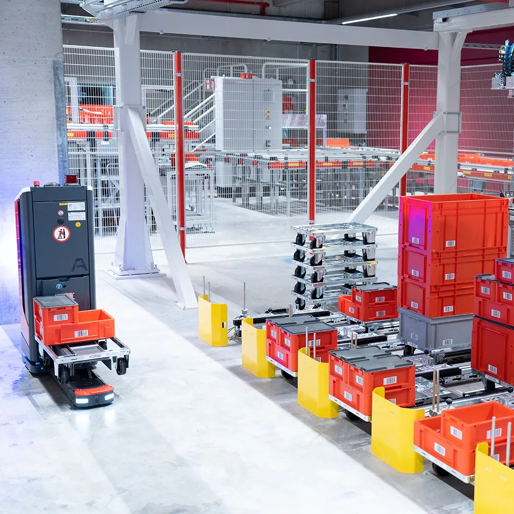 Automated warehouse area with mobile robot transporting red plastic crates on metal racks and conveyor systems