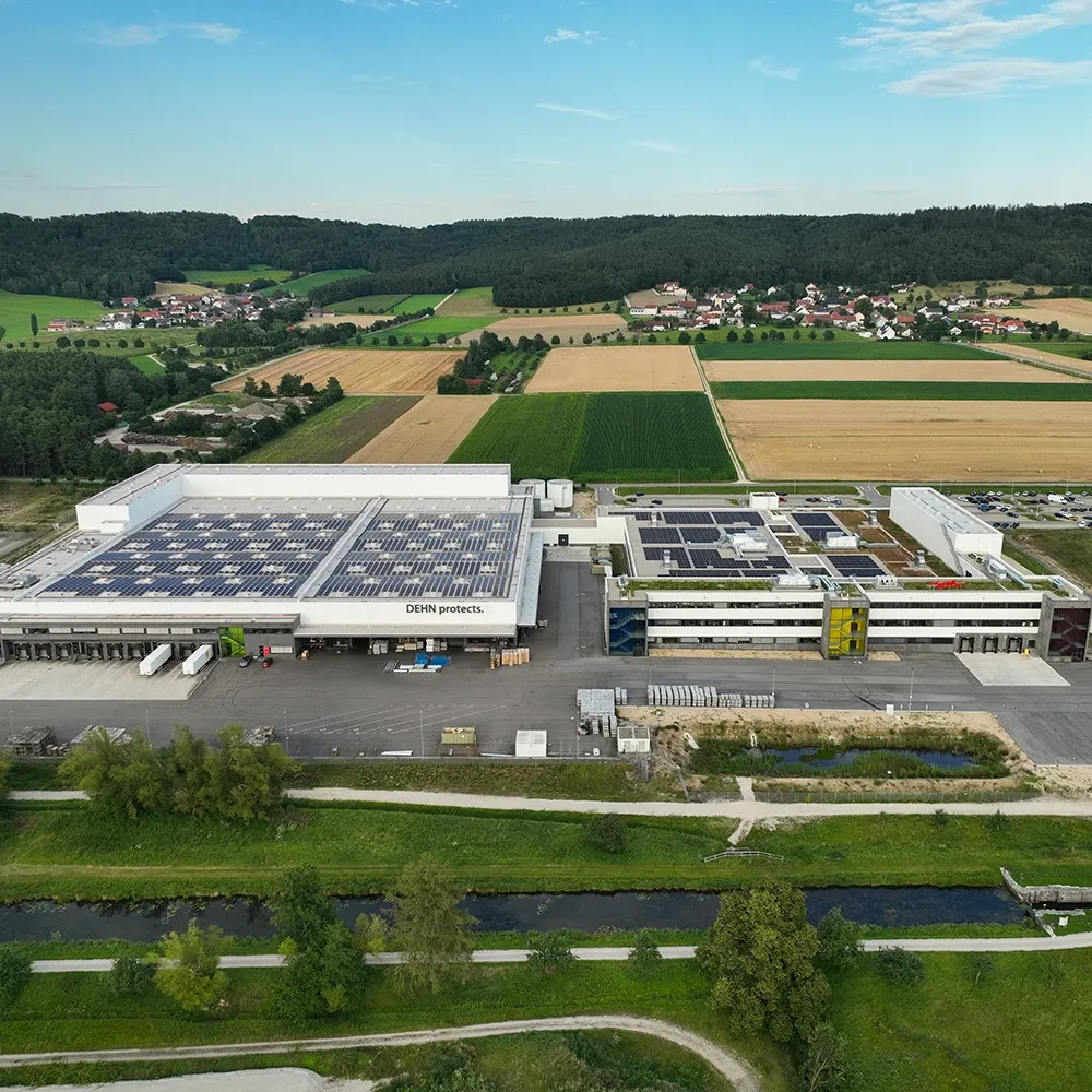 Aerial view of an industrial site with large warehouse buildings covered in solar panels, surrounded by fields and a small river.