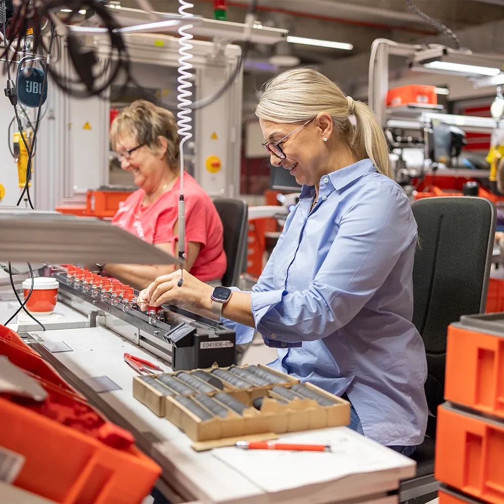Two women assembling electronic components at a workstation in a production facility, surrounded by tools and orange storage bins.