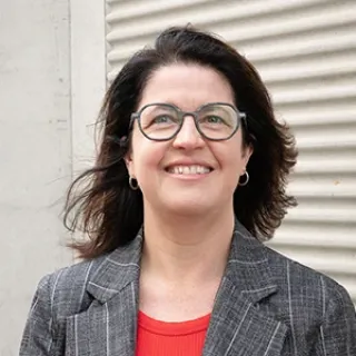 Portrait of a smiling woman wearing glasses, a gray blazer, and a red top, standing in front of a light-colored wall.