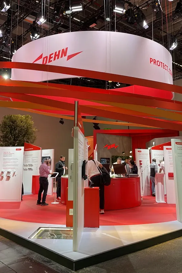Exhibition booth with red design and white-red Dehn logo on circular banner, several people viewing display panels and exhibits.
