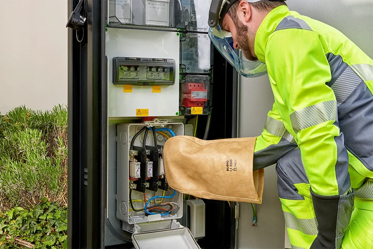 Technician in protective clothing working on an open electrical cabinet using insulated gloves for electrical safety.